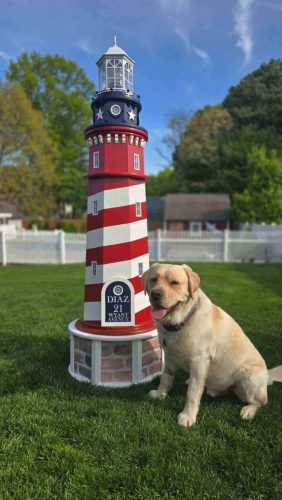 A yellow Labrador retriever sits on grass next to a red, white, and blue decorative lighthouse in a fenced backyard.