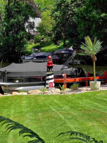 Two covered boats are docked on a calm river beside a small decorative lighthouse, with a well-kept lawn and potted palm tree in the foreground.