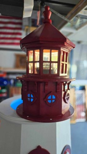 A close-up view of a red and white model lighthouse with lit windows, displayed indoors with an American flag in the background.